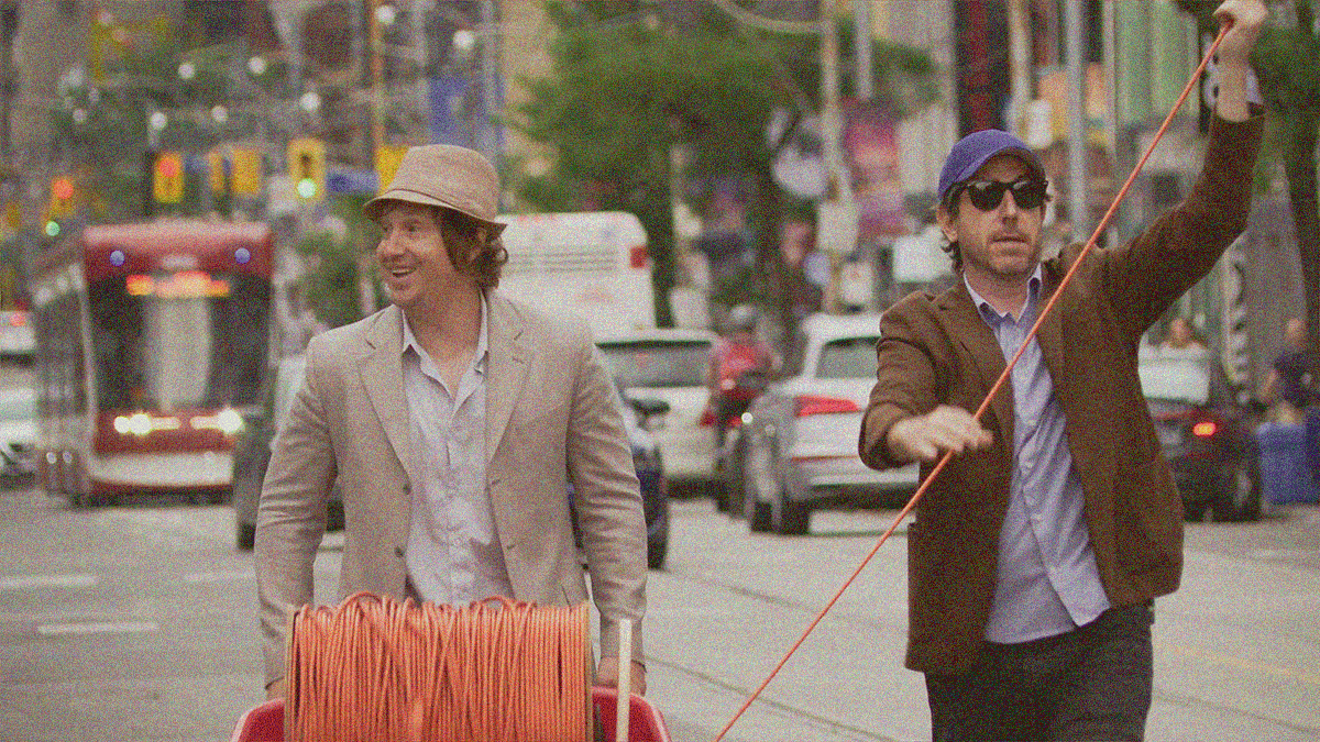 Two men walking down a Toronto street. The one on the left is rolling a wheelbarrow housing a giant spool of wire. The one on the right is carefully maintaining the integrity of said wire as it inexplicably shoots off into the sky through the top right corner.
