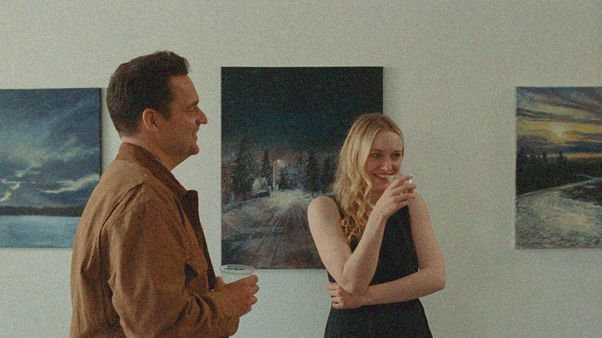 A man and a woman laugh with drinks in-hand at a gallery with paintings on the wall.