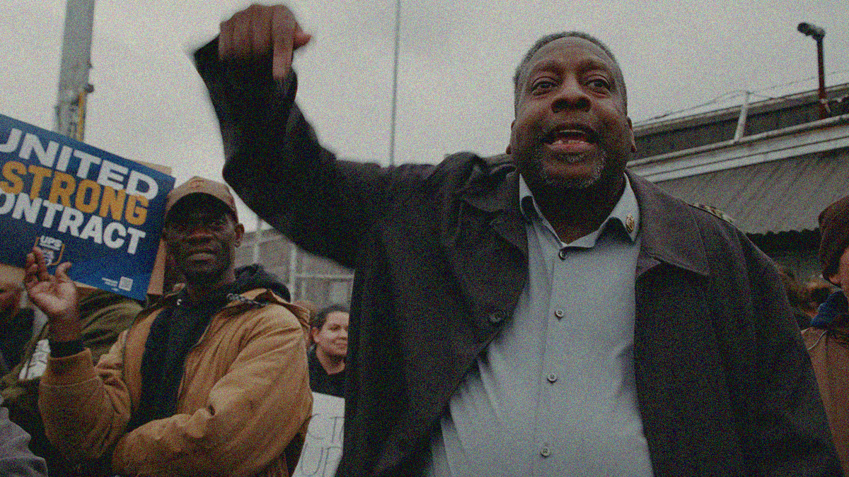 A man emphatically points while shouting in the streets. A crowd with a sign the says "United Strong Contract" is seen behind him.