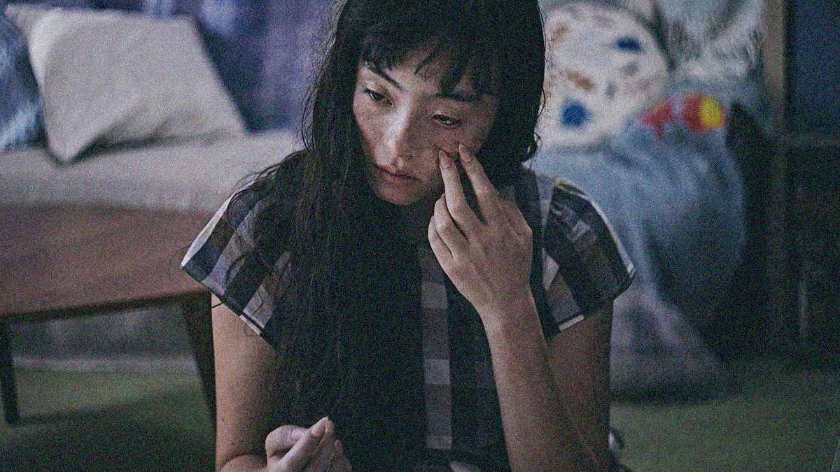 A woman is sitting on the floor. We see her from the waist up, head tilted and two fingers with razor sharp nails touching a bloodied cheek.