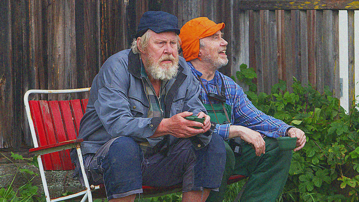 Two older men in hats sit outdoors on a bench with cups in-hand.