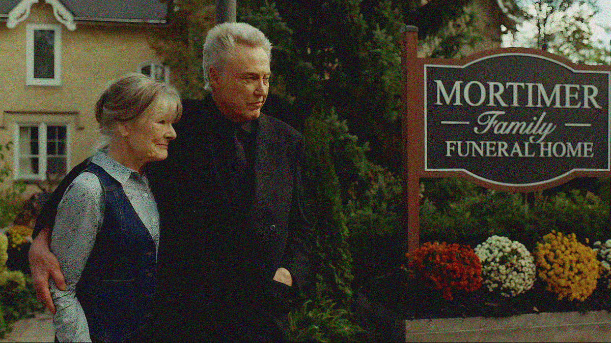 An older couple stands with arms around each other outside a family funeral home, both smiling and looking off-screen right.