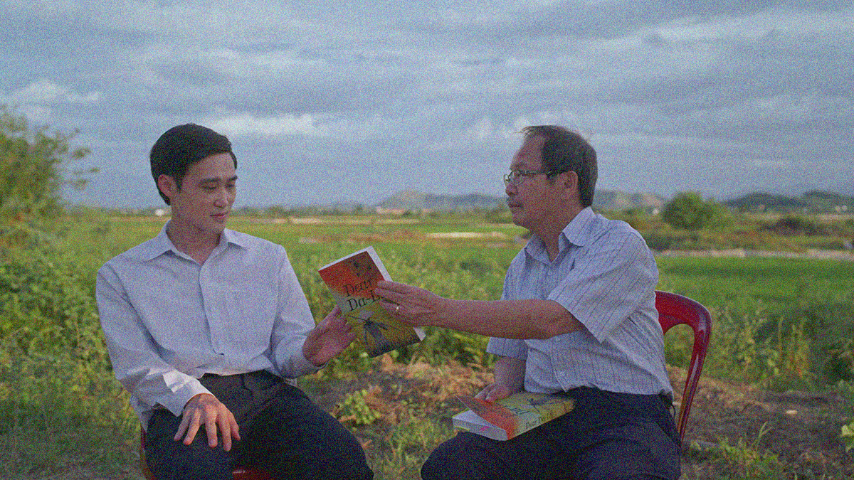 Two men sitting outside. The older on on the right hands the younger one on the left a book.