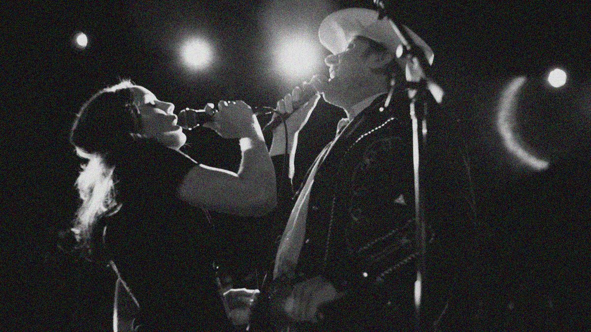 Black and white shot from below of a woman and man facing each other on-stage singing.