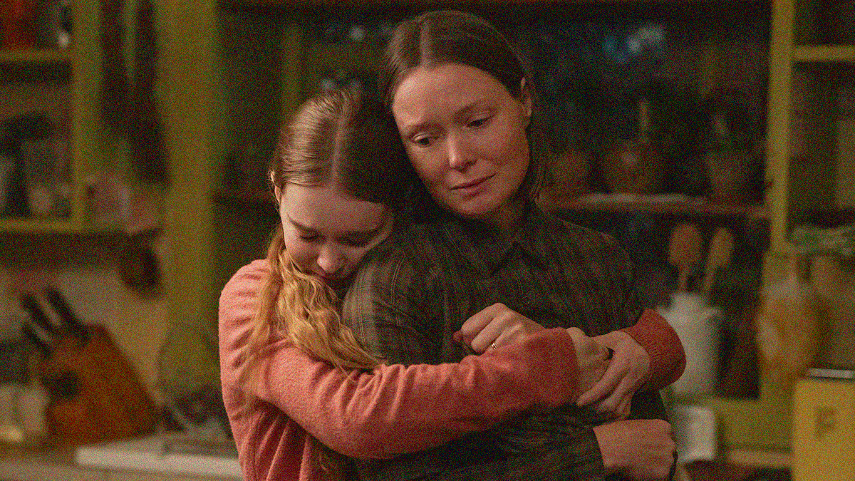 A teen stands behind her mother and wraps her hands around her chest in an embrace.
