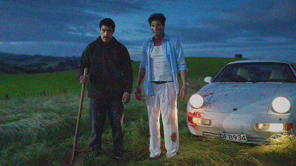 Two men the worse for wear are standing in a field beside a dirty white Porsche. The one holds a shovel.