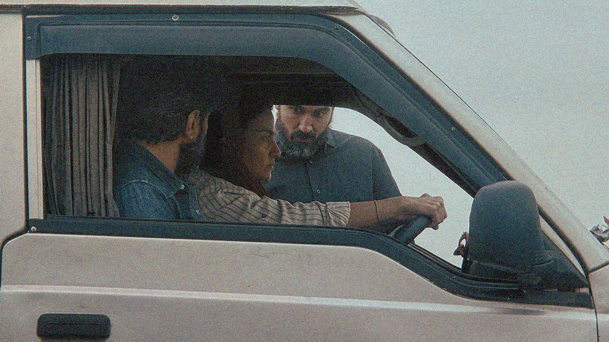 Image of the front seat of a van from outside the passenger side window. The man in the passenger seat is turned away to look at a man standing outside the vehicle and glaring at the driver. She sits with a determined stare out the front window with both hands on the wheel.