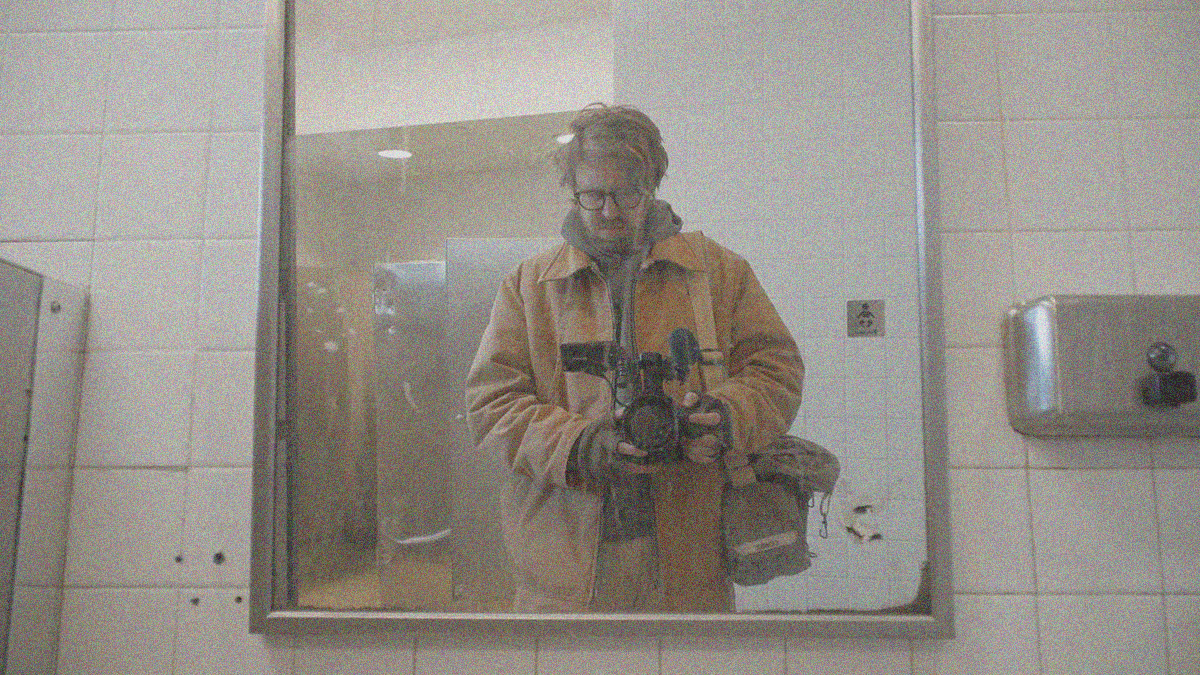 The reflection of a man holding a camera through a public bathroom mirror.