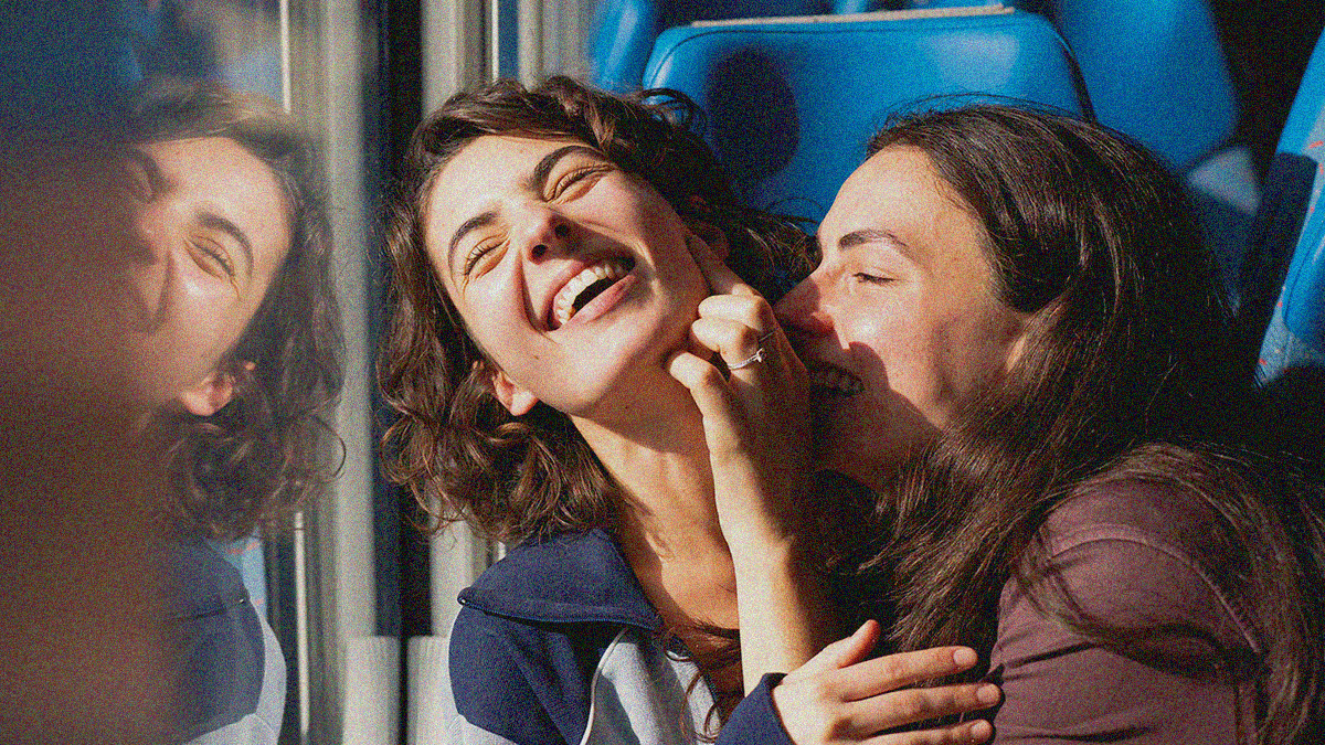 Close-up of two young women laughing together on a train. The one's hand is on the other's face.
