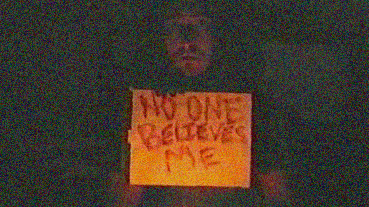 Grainy image of a man holding a yellow lined pad of paper up with the words "No one believes me" written in blood.