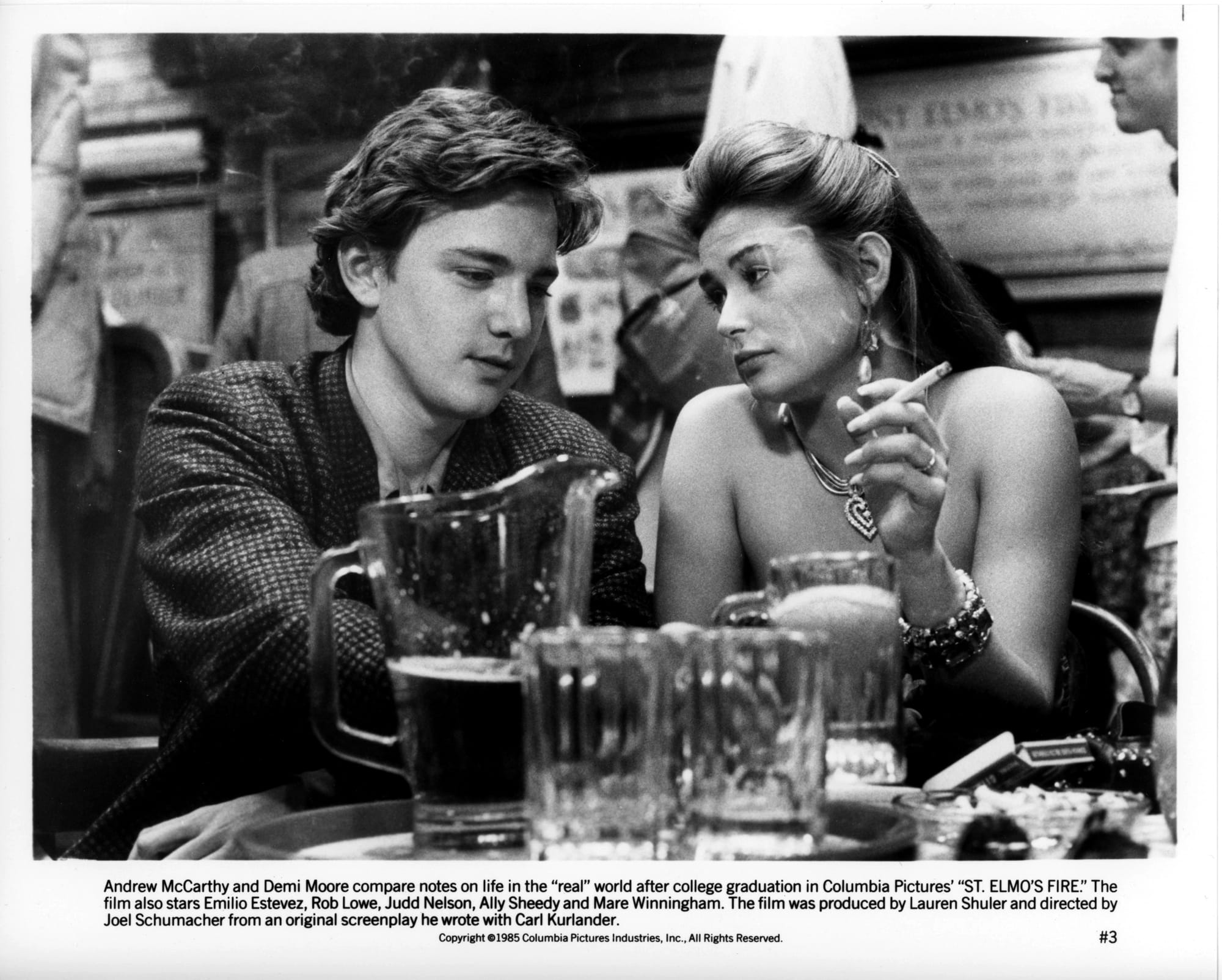 A young man and woman sit at a restaurant table. She has a lit cigarette in her hand. A pitcher of beer and glasses sit between them and us.