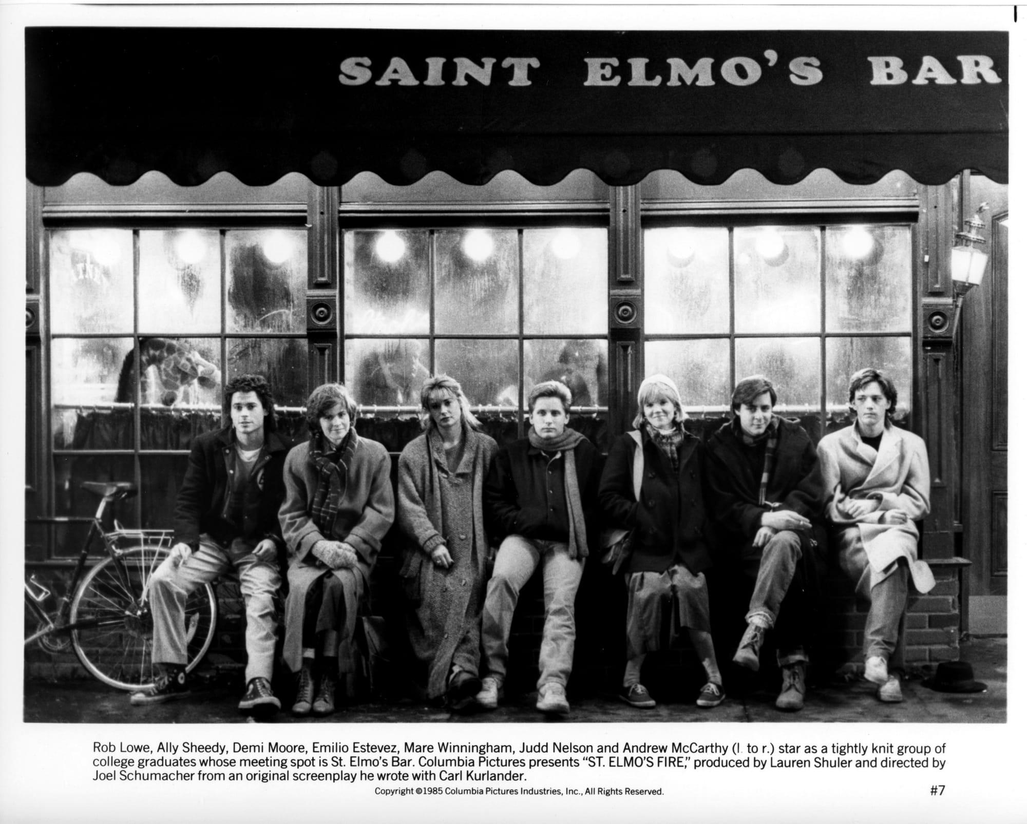 A group of seven young men and women sit side by side on the outdoor ledge of a bar window. The words SAINT ELMO'S BAR is seen on the awning above them.