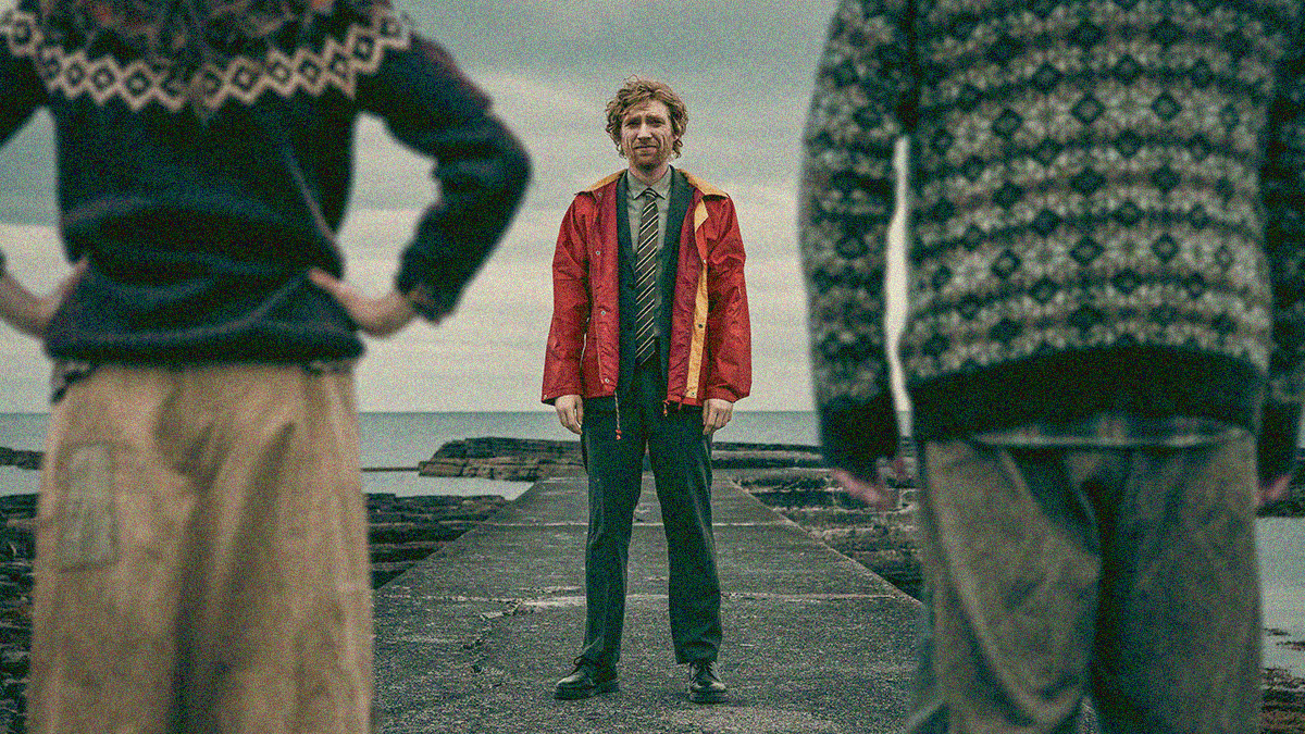 A man standing on a pier in an orange coat is seen looking towards the camera between the backs of two figures facing him in the foreground.
