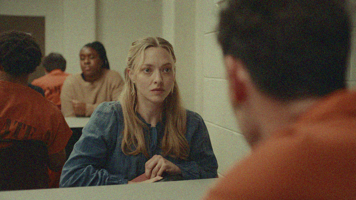 A woman with long blonde hair is seated and staring at a blurred man wearing an orange jumpsuit who's seated opposite her in the foreground.