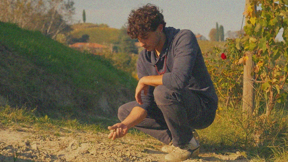 A young man crouches on dry vineyard soil with a clump of dirt in his hands.