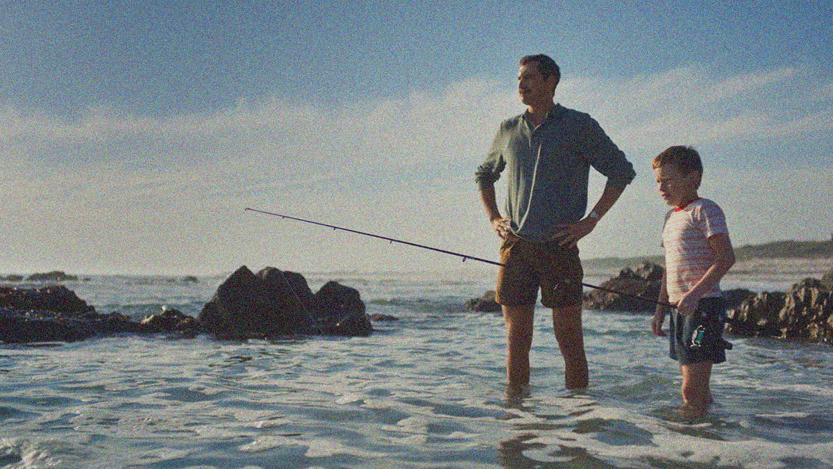A father stands with hands on hips beside his crying young son in choppy water. The latter holds a fishing rod in his hand.