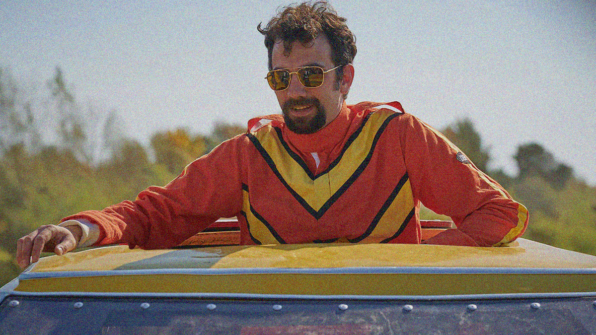 A goateed man wearing sunglasses in an orange and yellow jumpsuit is popped out of a car's sunroof.
