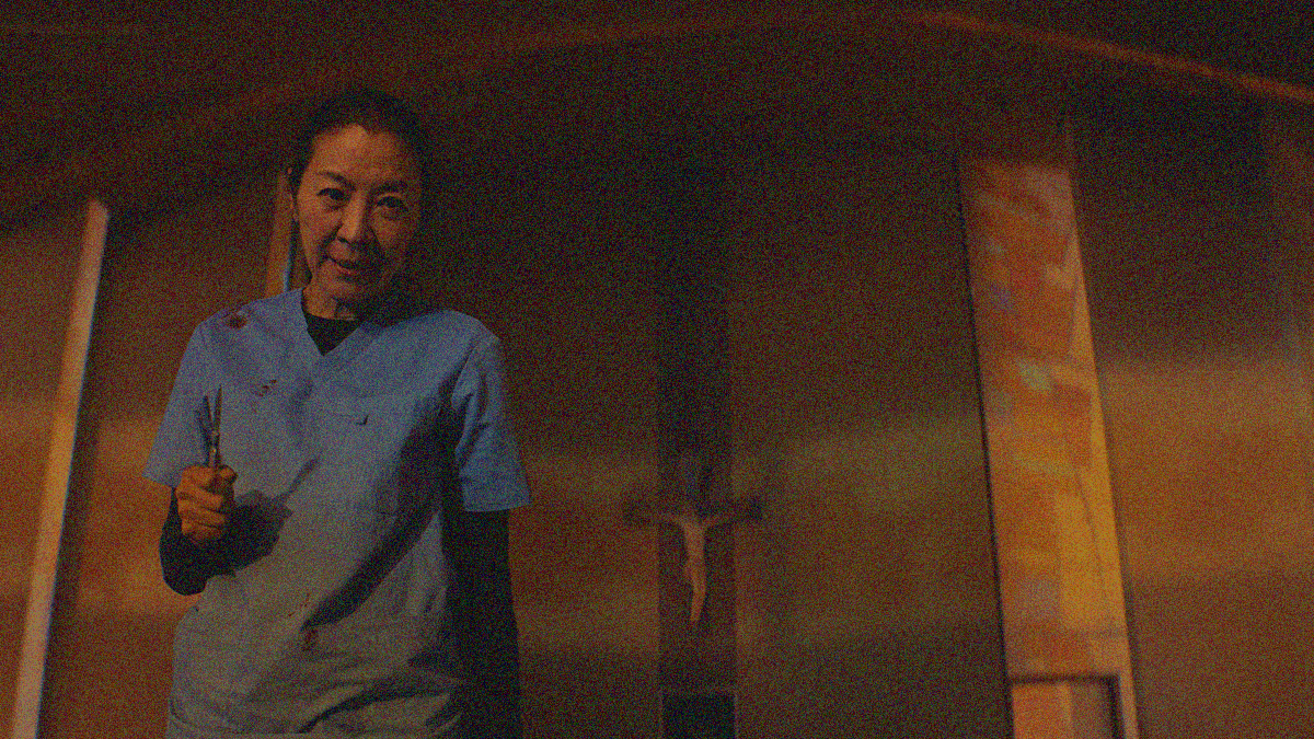 A woman in bloody blue scrubs looks downwards menacingly while holding a scalpel. A crucifix is on the wall behind her.