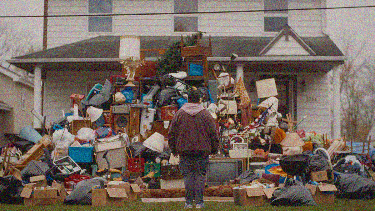 A shot from behind a man standing in front of and looking at a giant mound of trash piled on the lawn. A white house is behind it.
