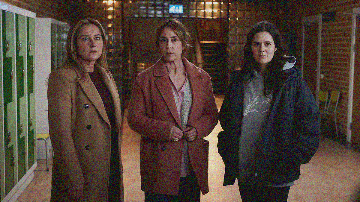 Three women in coats stand side by side looking towards the camera in a school hallway.