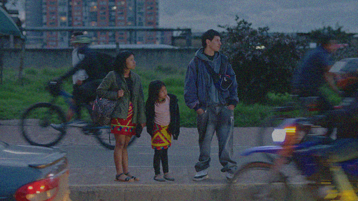 A woman holding the hand of her young daughter and a man stand on the curb of a street as a car and motorcycles drive by.