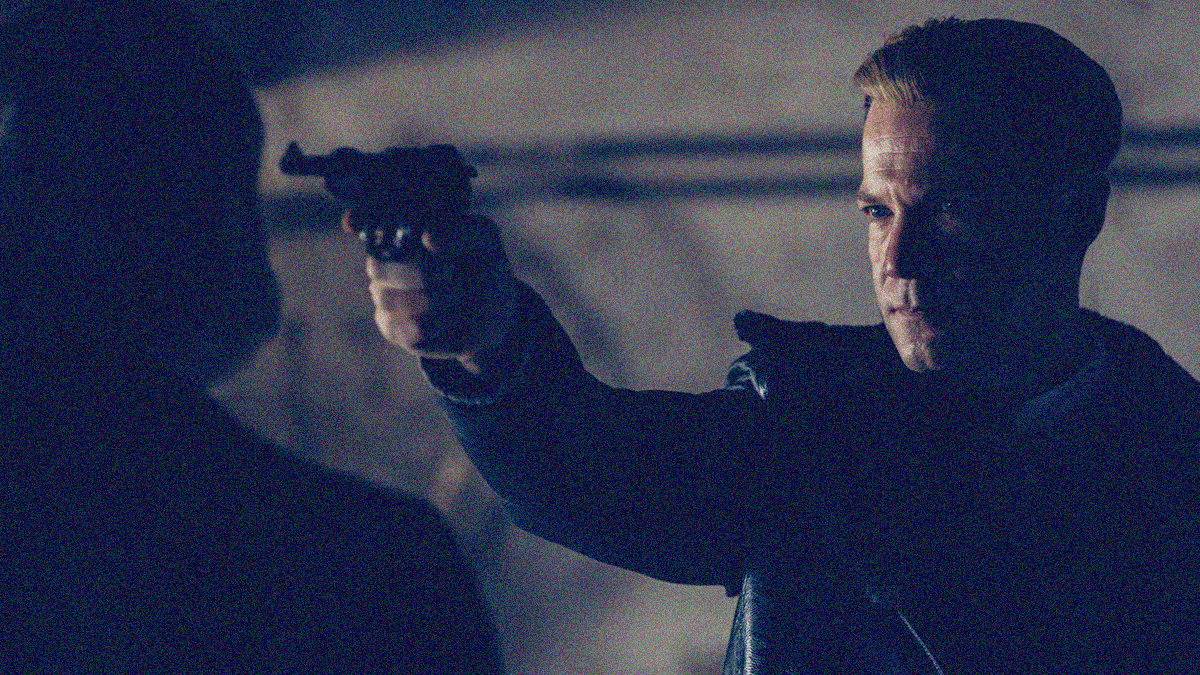 A man holds a Luger up to the face of another man in the dark.