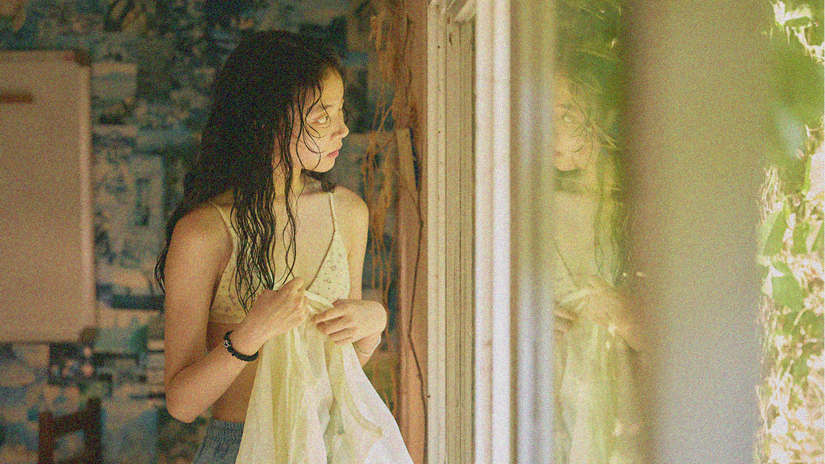 A teen girl in bra with wet hair gazes out a window while holding her shirt in her hands.