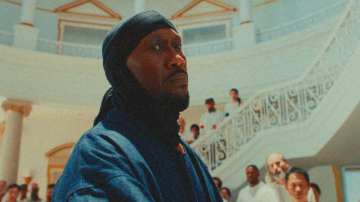 Close-up of a Black man staring menacingly off-camera with an ornate white staircase behind him filled with a bunch of people wearing white.