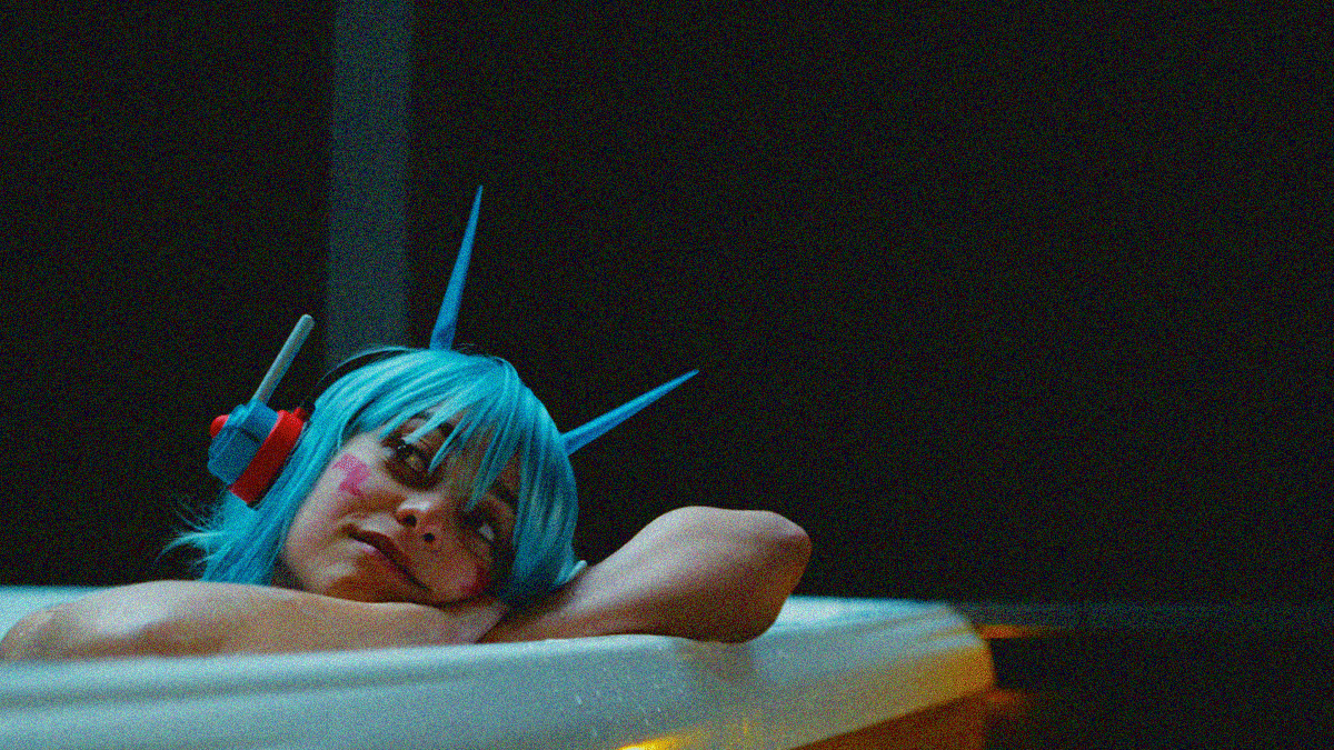 Close-up of a woman with blue hair and a headphones resting her head on her arms as she leans on the edge of a hot tub at night.