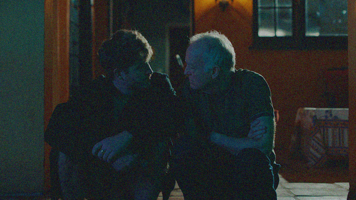 A young man (left) and older man (right) sit closely on a darkened porch at night, intently looking at the other.