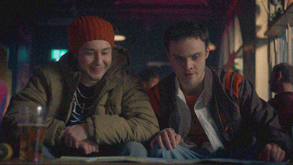 Two young men are seated, smiling, and looking at something on the table in front of them.