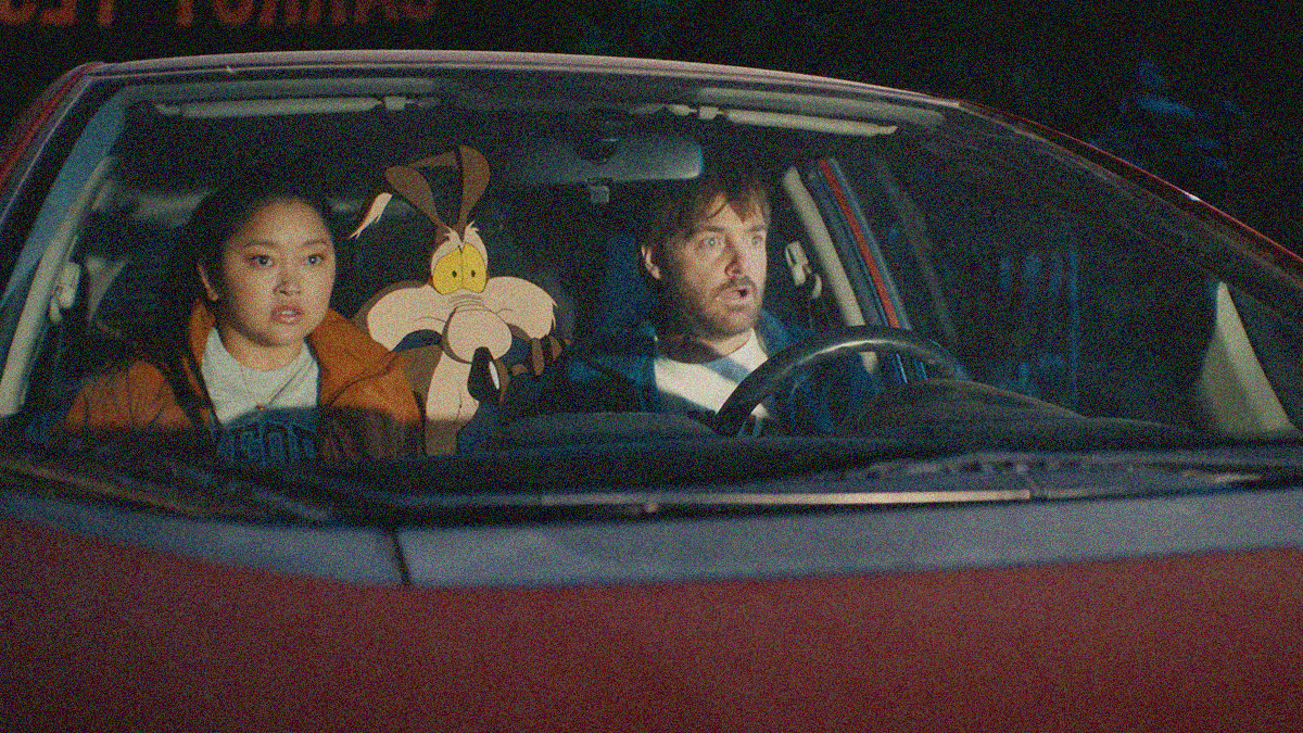 View inside a car from its hood that shows a young woman in the passenger seat and a man in the driver's seat with a cartoon coyote between them sitting in the back. All are wide-eyed looking at something off-screen right.