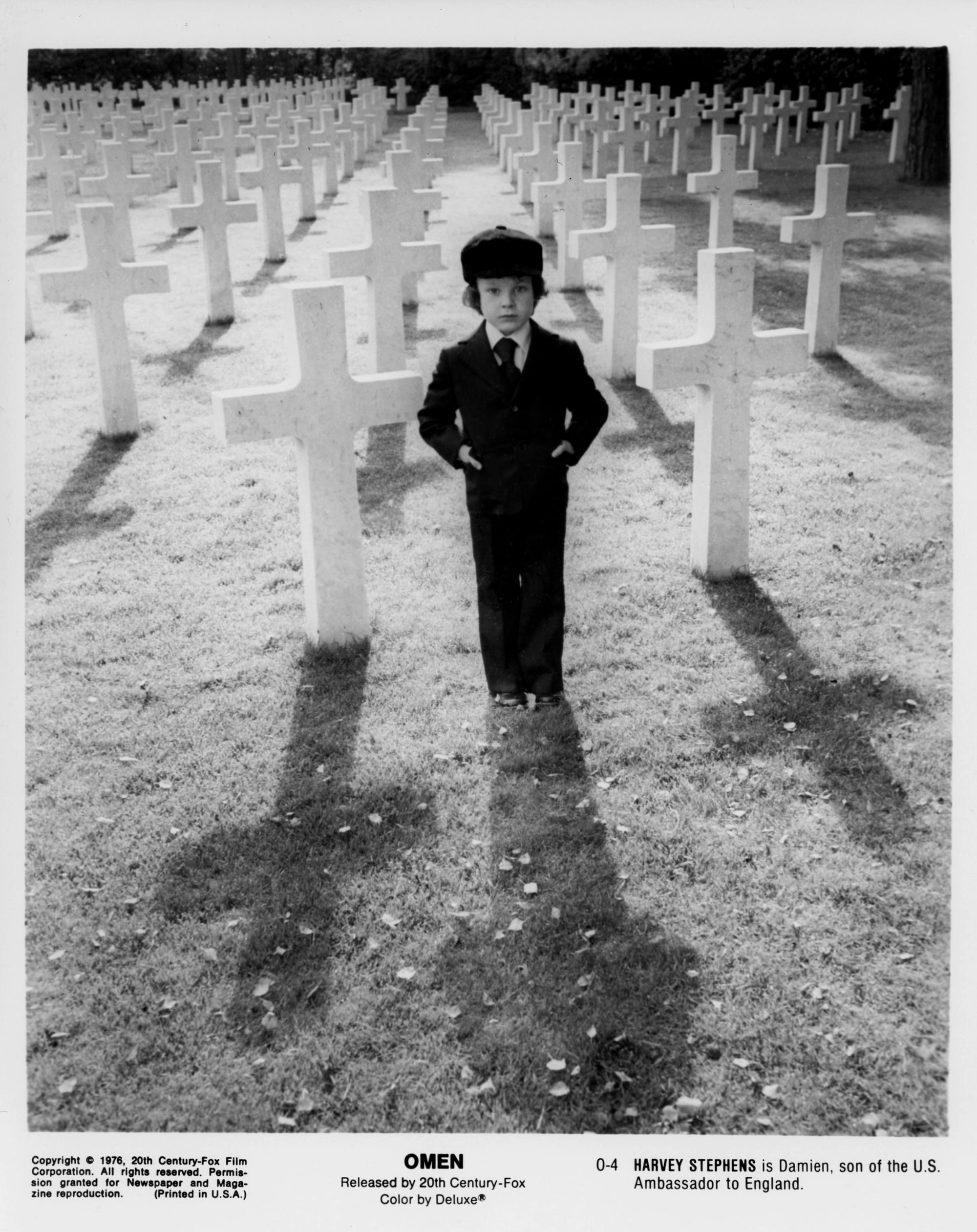 A young boy in black suit stand amongst rows of white crosses in a cemetary.