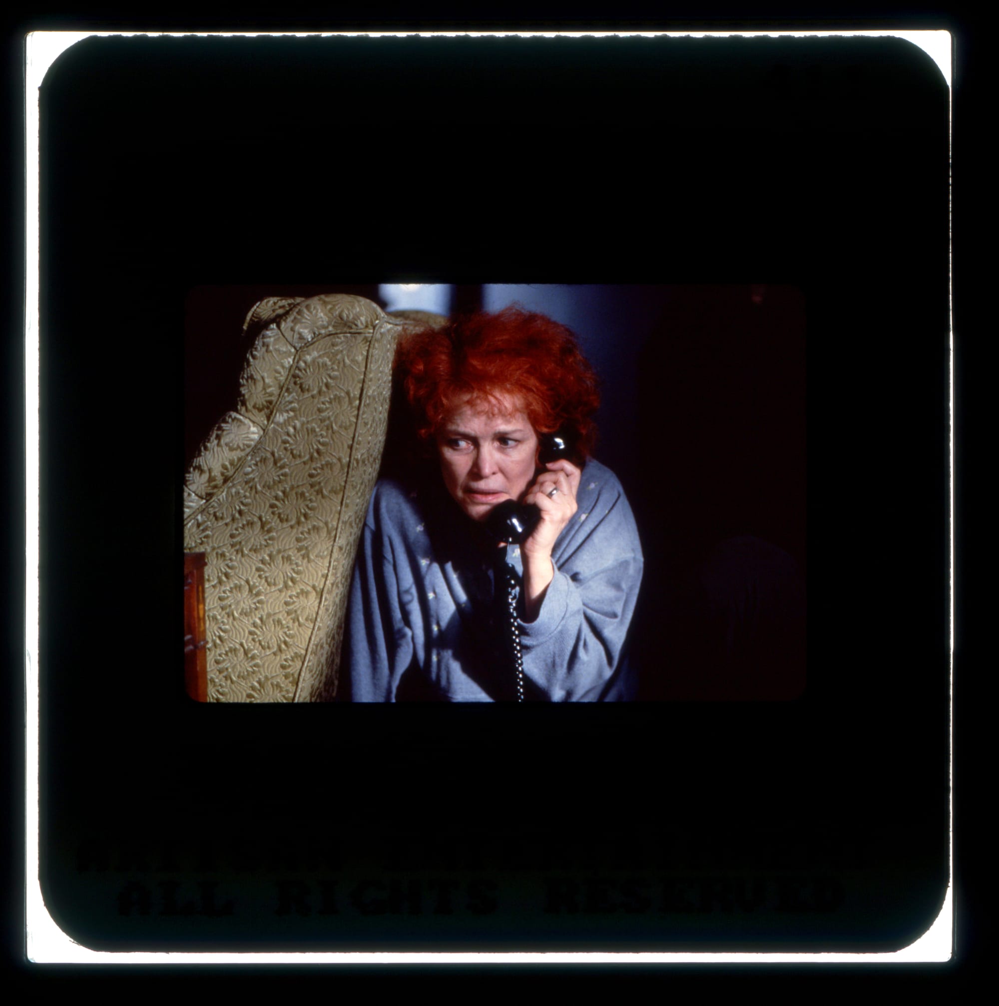 A scared woman in blue crouches behind an upholstered chair with a black phone receiver against her ear.