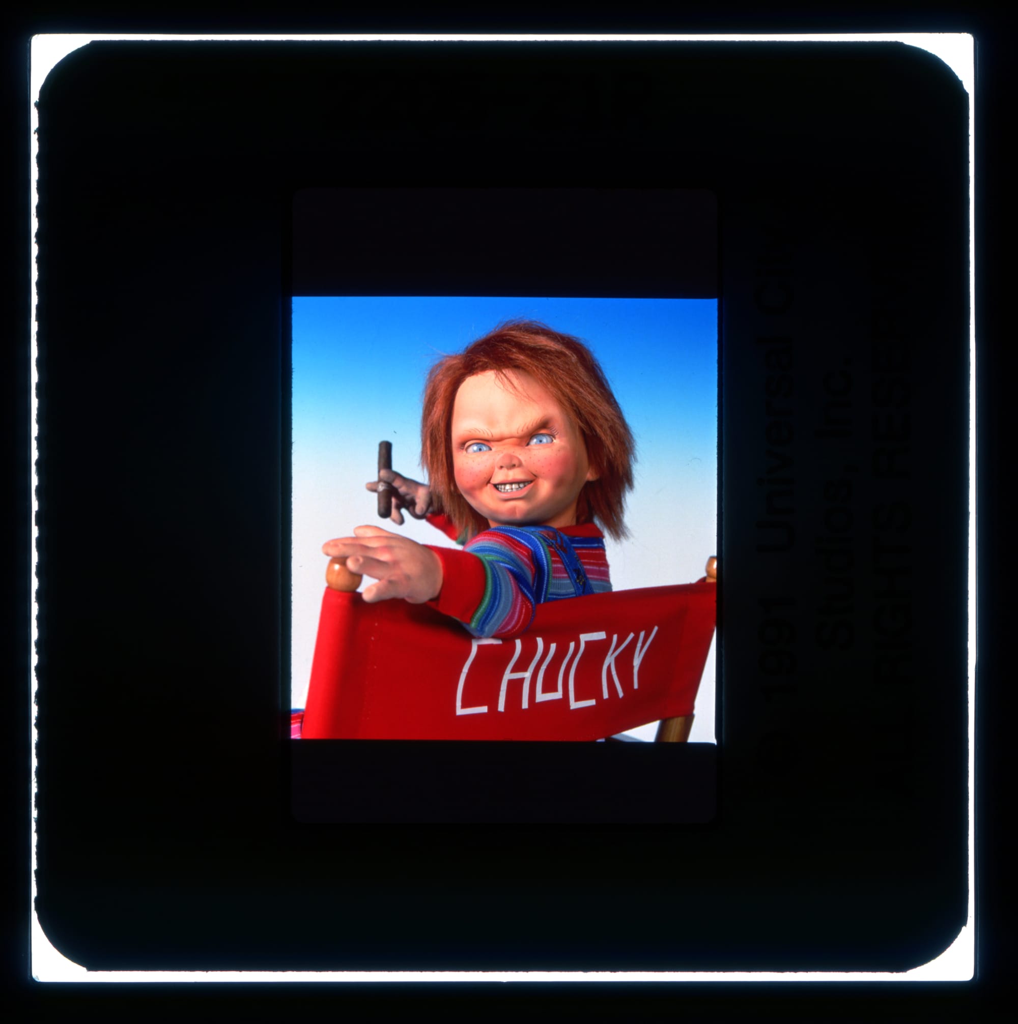 Portrait of the Chucky doll sitting in a director chair from behind. He's turned to look at us over the red backer with his name written in white tape. He smiles with a cigar in hand.