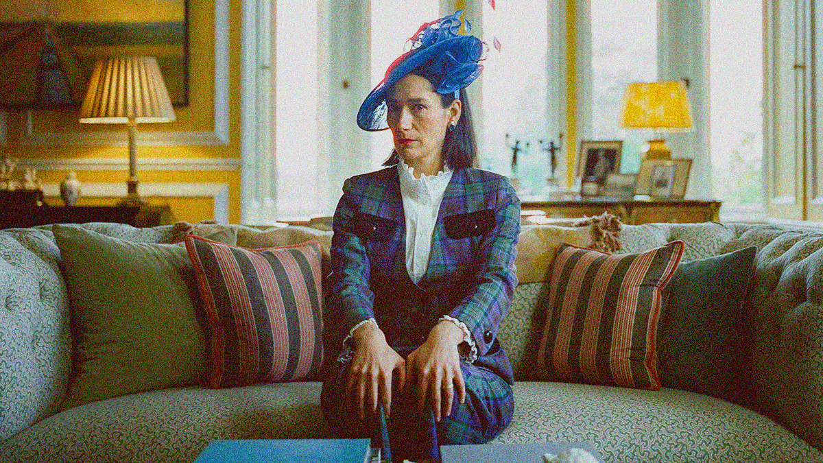 A woman in a pants suit with a blue hat sits on the center of a couch with hands posed on knees.