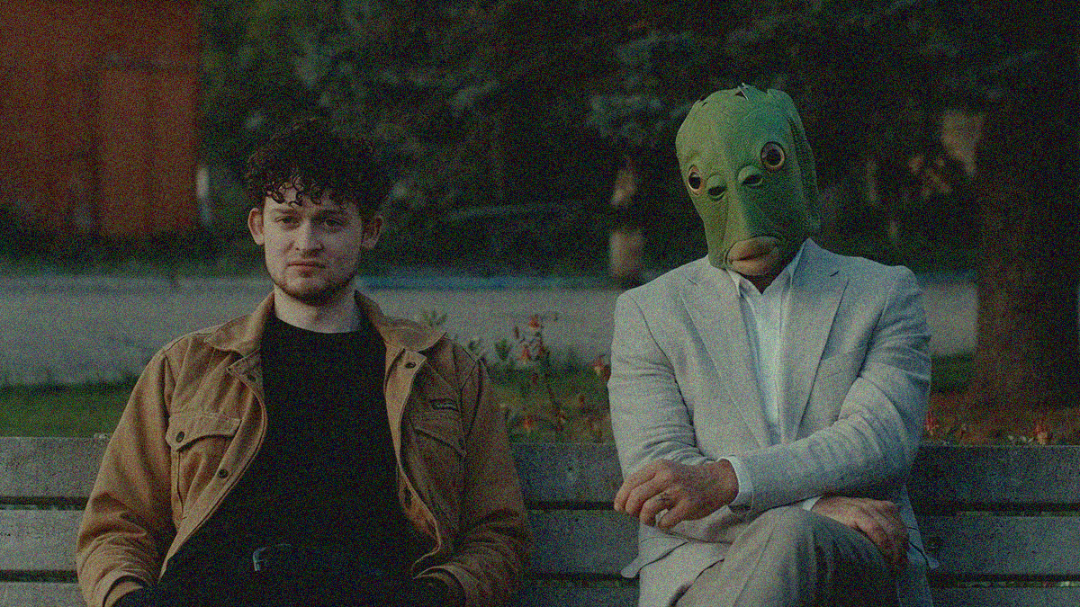 Close-up of two men sitting on a park bench—the one on the right is wearing a rubber fish mask.