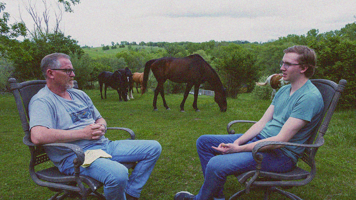 An older father in a chair on the left listens as his son speaks from a chair on the right. They are outside and horses can be seen grazing between them.