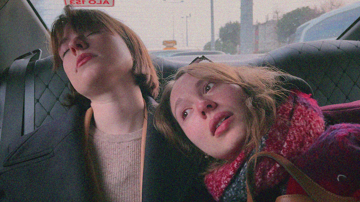 Two women sit in the back of a taxi. The one has her eyes closed, the other rests her head on the former's shoulder.