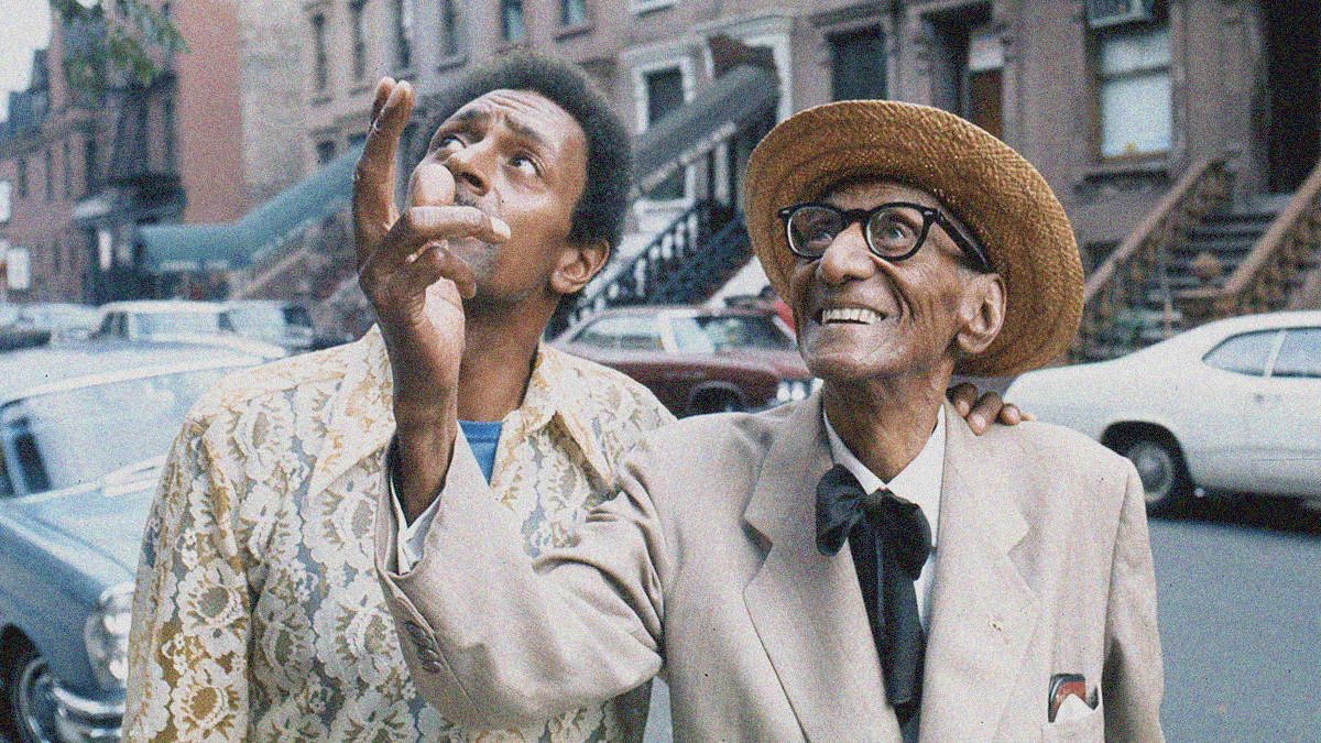 An old Black man in a suit and hat stands next to a younger man while smiling and lifting his right arm upwards while on a Harlem street.