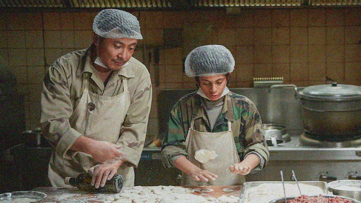 A man and a teen wearing aprons and hairnets stand behind a table covered in dough and flour.