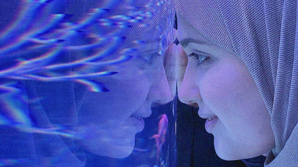 Close-up of a young woman with headscarf leaning her forehead against a glass fish tank so her reflection can be seen smiling back.