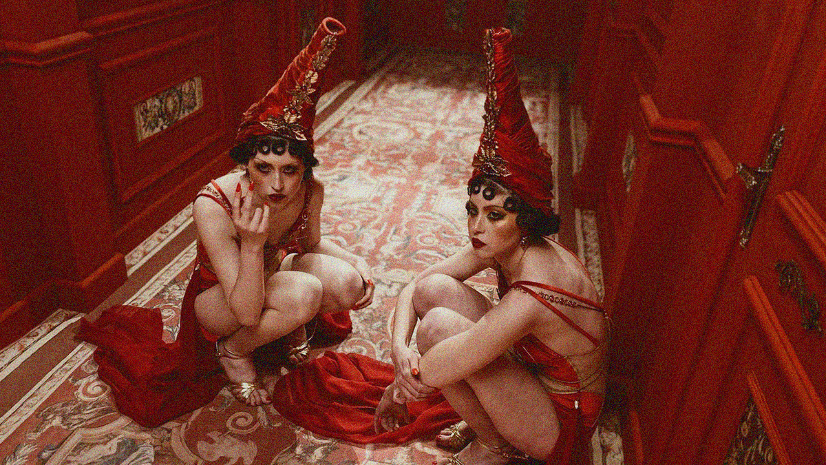 Two women in red dresses with large red conical hats crouch on the floor of a hallway of red doors.
