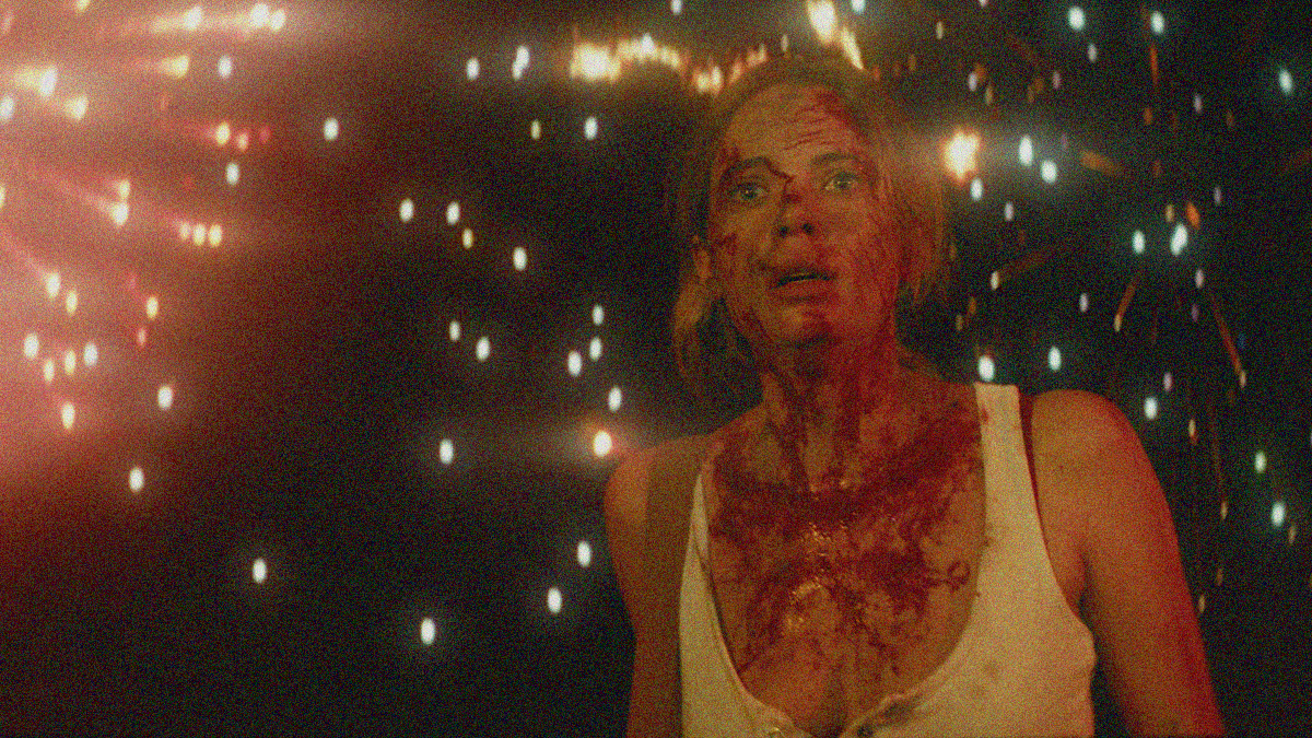 A woman in a buttoned tank top covered in blood while fireworks light up the night sky behind her.