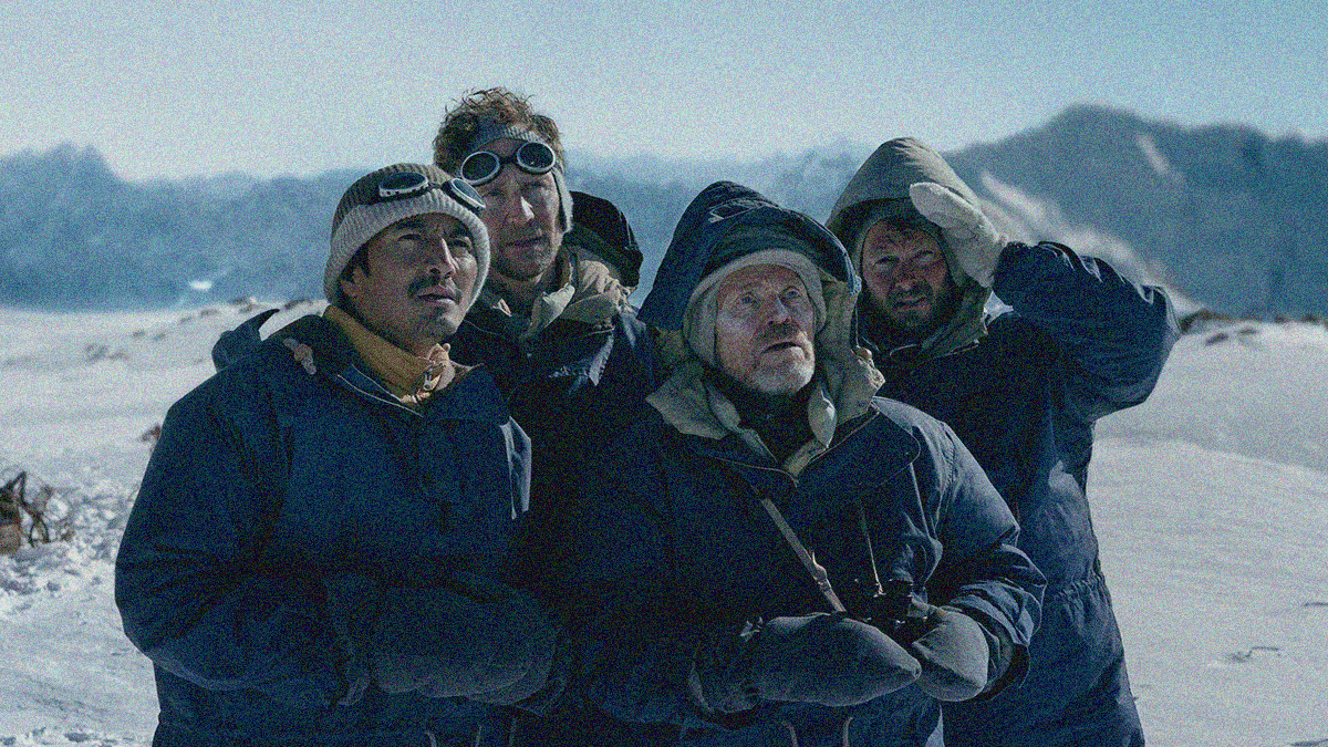Four men in winter coats and mittens in the snow against mountains who are all looking upwards off-screen right.