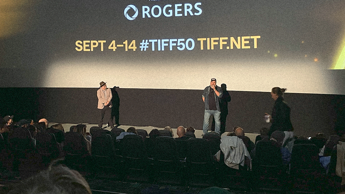 Two men stand before a movie theater audience with TIFF's branded info projected on screen behind them.
