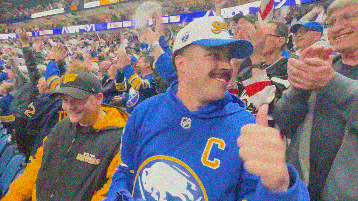 A man in Sabres apparel looks off-screen right with a smile and thumbs up while a man in Bruins apparel laughs to his right.