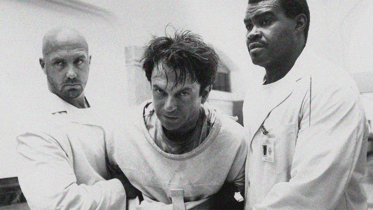 Sam Neill in straitjacket (flanked by two orderlies carrying him off-screen left) stares directly into the camera.