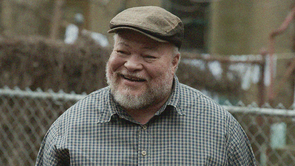 A genial bearded man smiles in a flat cap with a chain link fence seen behind him.