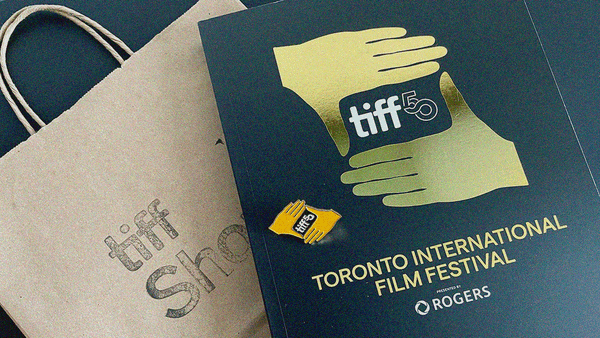 A brown paper bag with "TIFF Shop" stamped on it; TIFF 50 program book and pin with logo between two hands making a viewfinder.
